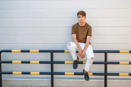 年轻英俊的男子，与一个发型在时尚夏天的衣服，坐在靠近现代的有光泽的灰色墙体