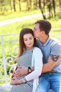 英俊的男人和他美丽的怀孕的妻子是拥抱，微笑着站在公园