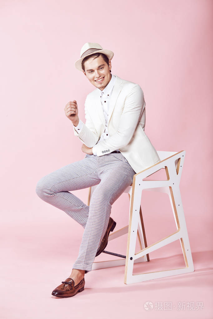 年轻美丽微笑男模特穿着白色夹克 白衬衫 灰色的裤子和白色的帽子，坐在椅子上在工作室与浅粉红色背景