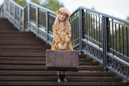 甜美的女孩在一个手提箱站在楼梯上等待上一顶帽子。