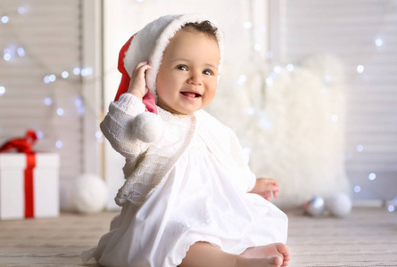 可爱的小婴儿在圣诞老人的帽子坐在地板上对模糊的圣诞灯