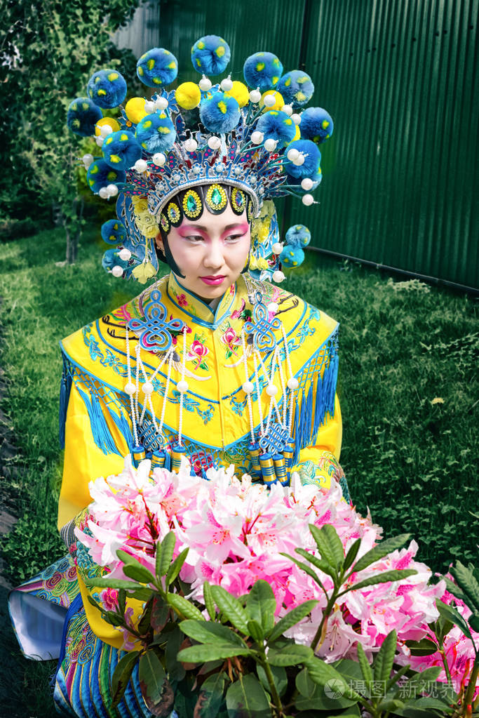 美丽的中国女孩，在传统蓝色和黄色中国服饰和绒球帽子坐在不远处的粉红色的花灌木