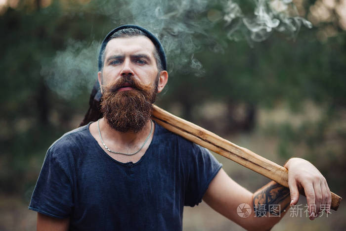 一个时尚的樵夫,留着长胡子和时髦的衣服抽的香烟和树林的手里拿着一