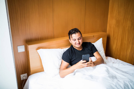 英俊的年轻男子，穿着睡衣躺在床上在早上使用一部手机
