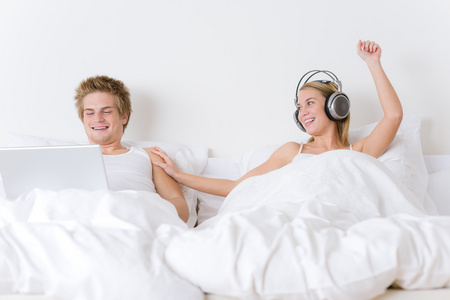 年轻夫妇放松躺在床上与音乐和便携式计算机