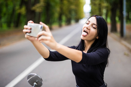 年轻漂亮的女人坐在滑板车自拍照以她的智能手机上的室外肖像