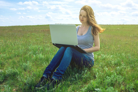 妇女坐在绿色的草地上, 天空的背景云和工作或学习与笔记本电脑无线