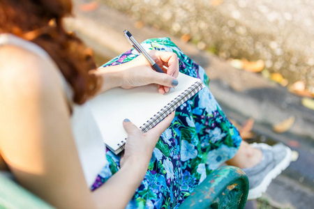 用钢笔写的笔记本，坐在木凳上，公园关闭了左手亚洲妇女