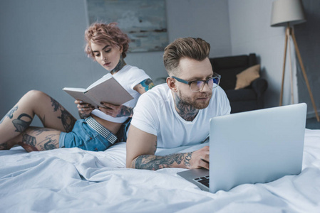 纹身女友看书, 而男友在床上使用笔记本电脑