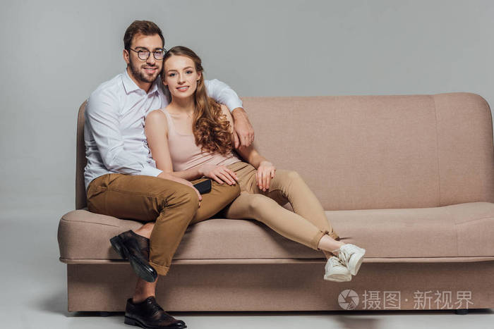 快乐时尚的年轻情侣与智能手机微笑在相机上, 而坐在一起在沙发上的灰