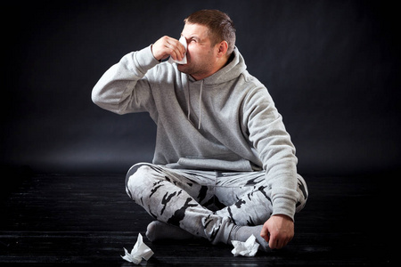 一个穿着运动服的年轻可怜的男人坐在地板上, 手里拿着餐巾, 在黑色的