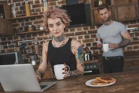 在早餐时使用笔记本电脑的纹身女孩, 在厨房的男朋友后面