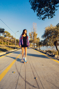 时髦的酷女孩在紫色夹克是骑在路上的一个长板