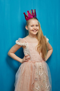 微笑的女孩穿着优雅的粉红色的礼服和纸冠在蓝色的墙壁背景, 圣诞节概念