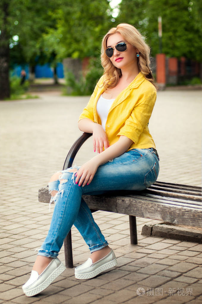 一个迷人的年轻的金发女郎时装模特坐在公园的长凳上黄色夹克戴墨镜