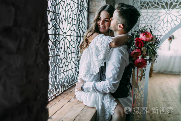 性格开朗的年轻新娘拥抱新郎虽然他吻她的脸颊上拥抱她的腰在窗台上