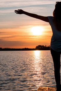 年轻女人站在木舟在湖上张开双臂，看着七彩的晚霞