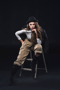 美丽的年轻女子在时尚的帽子和冬季夹克坐在梯子上, 看着在黑色的摄像头