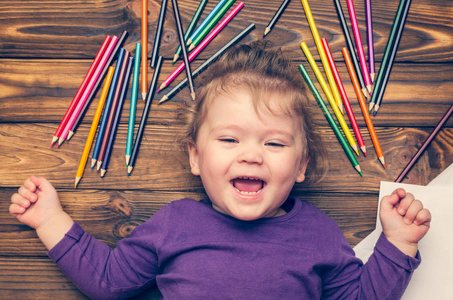 笑的小女孩躺在木地板上, 散落着彩色的铅笔