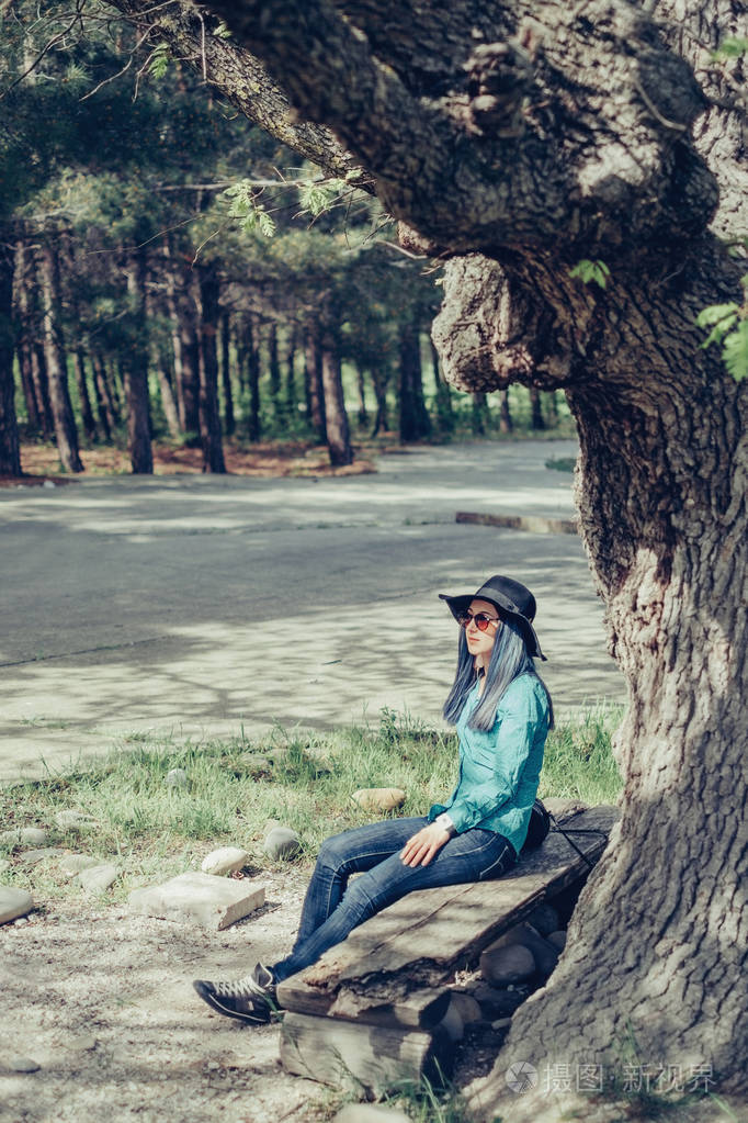 休闲风格的年轻女子在阳光明媚的日子在橡树上的长凳上休息