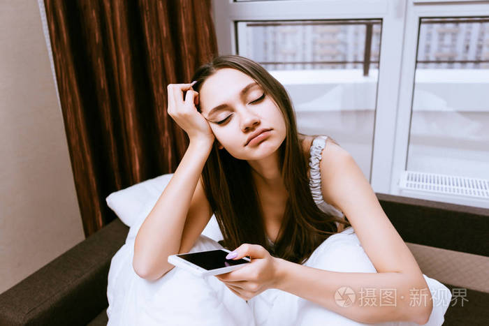 瞌睡的小女孩早早醒来, 坐在床上, 手里拿着智能手机, 想睡觉