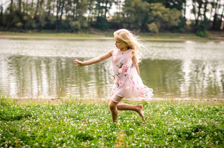 美丽的白种女孩与长金色的头发在粉红色的礼服跳舞作为仙女在绿草河边