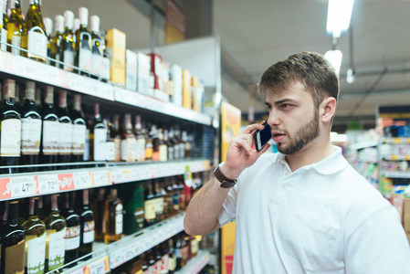 一个严肃的人在超级市场用电话讲话。一个男人在商店里选择酒精。买方在超级市场购买产品