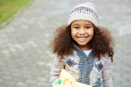 可爱的非洲裔美国女孩的画像抬头看着相机, 微笑着穿着针织帽和毛衣在温暖的秋日