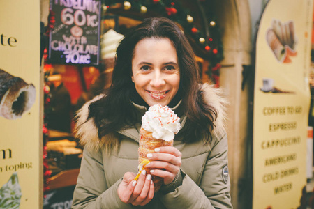 一个美丽的女孩在温暖的夹克吃 trdelnik 或 Trdlo 与奶油在她的手, 在冬天在捷克共和国, 布拉格在圣诞节市场