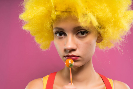 哀伤的女孩小丑在明亮的黄色假发吃 chupa chups, 被隔绝在粉红色背景