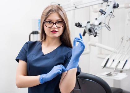 近现代牙科办公室美丽的女牙医的特写照片。医生戴着眼镜, 蓝色制服, 蓝色手套。牙科