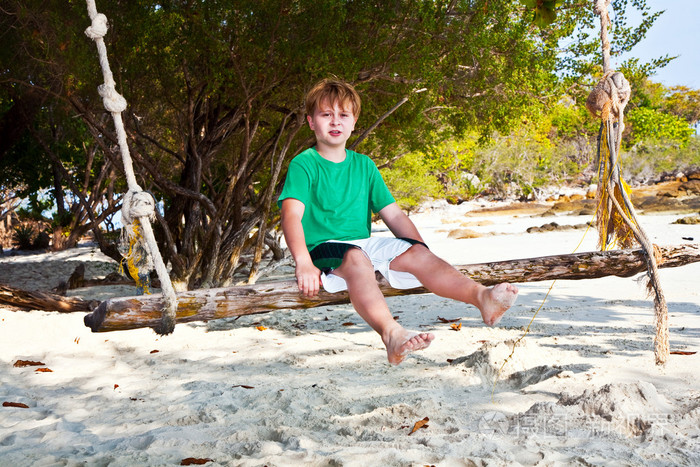 男孩坐在树下沙滩上的秋千上