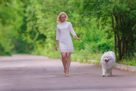 美丽年轻的金发女孩，穿着与夏天花园里的白色蓬松狗散步