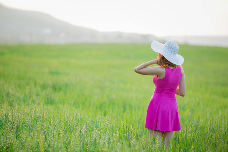 年轻女子穿着紫色的衣服和白色的帽子漫步在绿色的田野上, 背上有山