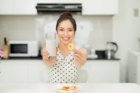 年轻的亚洲妇女拿着牛奶玻璃咬饼干在她的厨房