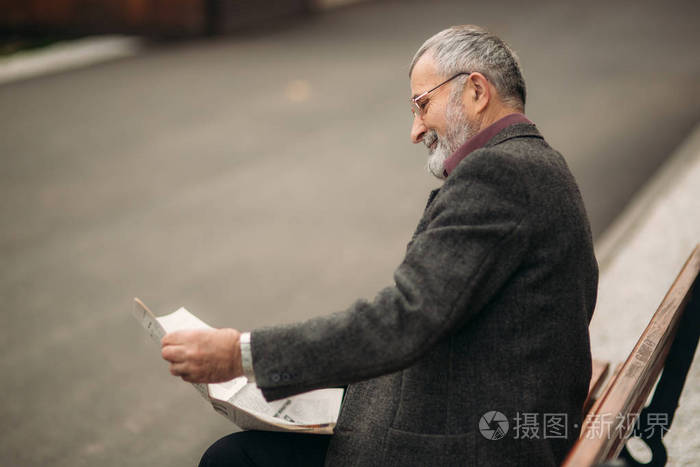 一个穿着灰色夹克的漂亮的祖父坐在公园的长椅上读报纸