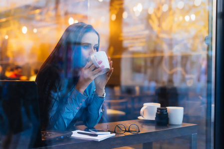 穿着蓝色衬衣的年轻女孩在咖啡馆里喝着香浓的咖啡, 看着窗外照片