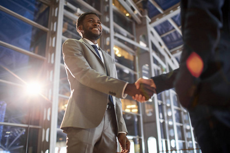 两个商业伙伴握手时的低角度肖像 微笑的非洲裔美国商人在现代玻璃办公楼大厅与白种同事握手