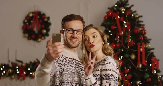 年轻快乐的情侣在爱拍漂亮的照片, 在一个智能手机和摆在圣诞气氛客厅。Selfies 的圣诞树背景。室内