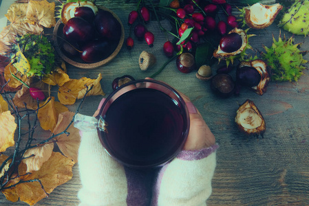 女性手捧着玫瑰花茶的茶杯, 捧着玫瑰花的臀部和栗子, 在木制背景下的秋叶。季节性的维他命饮料秋季背景