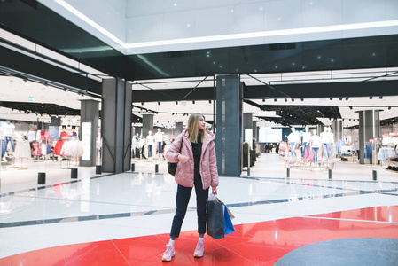 一个时髦的金发女郎, 手里拿着购物袋, 站在商场里, 面对着一家服装店的背景。