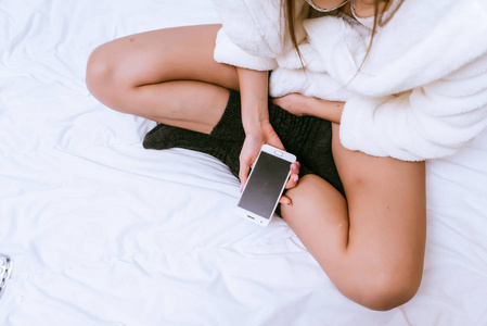 女孩坐在床上的白色大衣, 手持智能手机在她的手