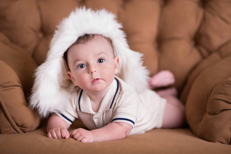 快乐的新生婴儿男孩粉红色的脸颊摆在格律复古休闲风格棕色沙发沙发与大白色毛皮冬季温暖的帽子