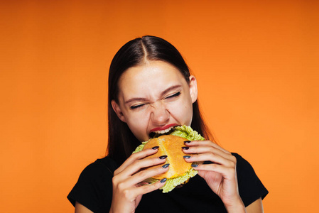 这个女孩急切地吃美味的汉堡。饿了的女孩吃。橘黄色的背景上孤立