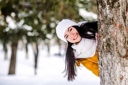 美丽的女孩在雪躲在一棵树后面的肖像