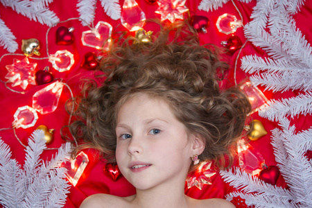 一个小 fairhaired 的女孩躺在红色的地板上, 圣诞花环和冷杉树枝围绕着她的头