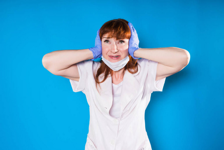 一个愤怒的护士戴着口罩和手套, 用手捂住耳朵, 看着摄像头, 在蓝色背景下隔离