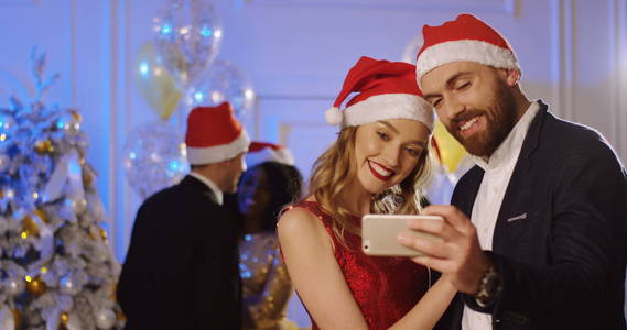 年轻幸福的情侣在圣诞老人的肖像在新年派对上用智能手机制作漂亮的 selfies。跳舞的人背景。室内