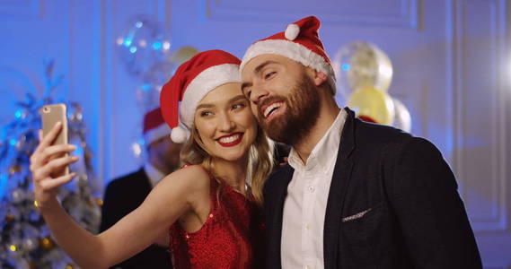 在圣诞晚会上, 一对年轻迷人的情侣在圣诞老人的帽子上做着滑稽的 selfies。跳舞的人背景。室内