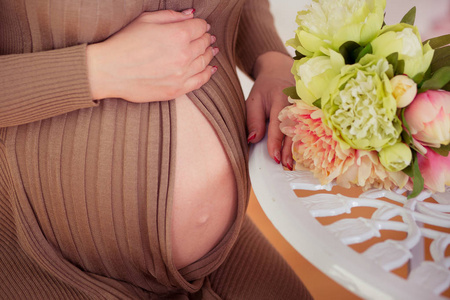 怀孕的女人摆在桌子旁边, 捧着一束开着棕色时髦连衣裙的鲜花, 抱着她腹中未出生的婴儿。腹部与大肚脐肚脐